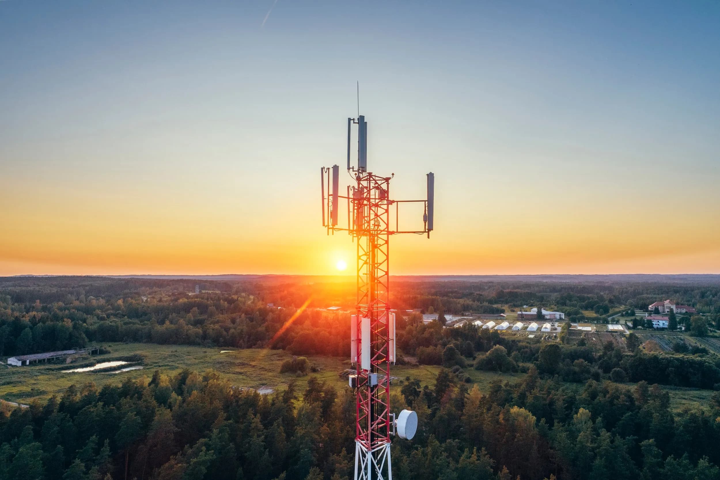 An aerial drone view looking at a lattice telecommunication cellular tower structure with the sun setting brightly over a green forest landscape in the background.