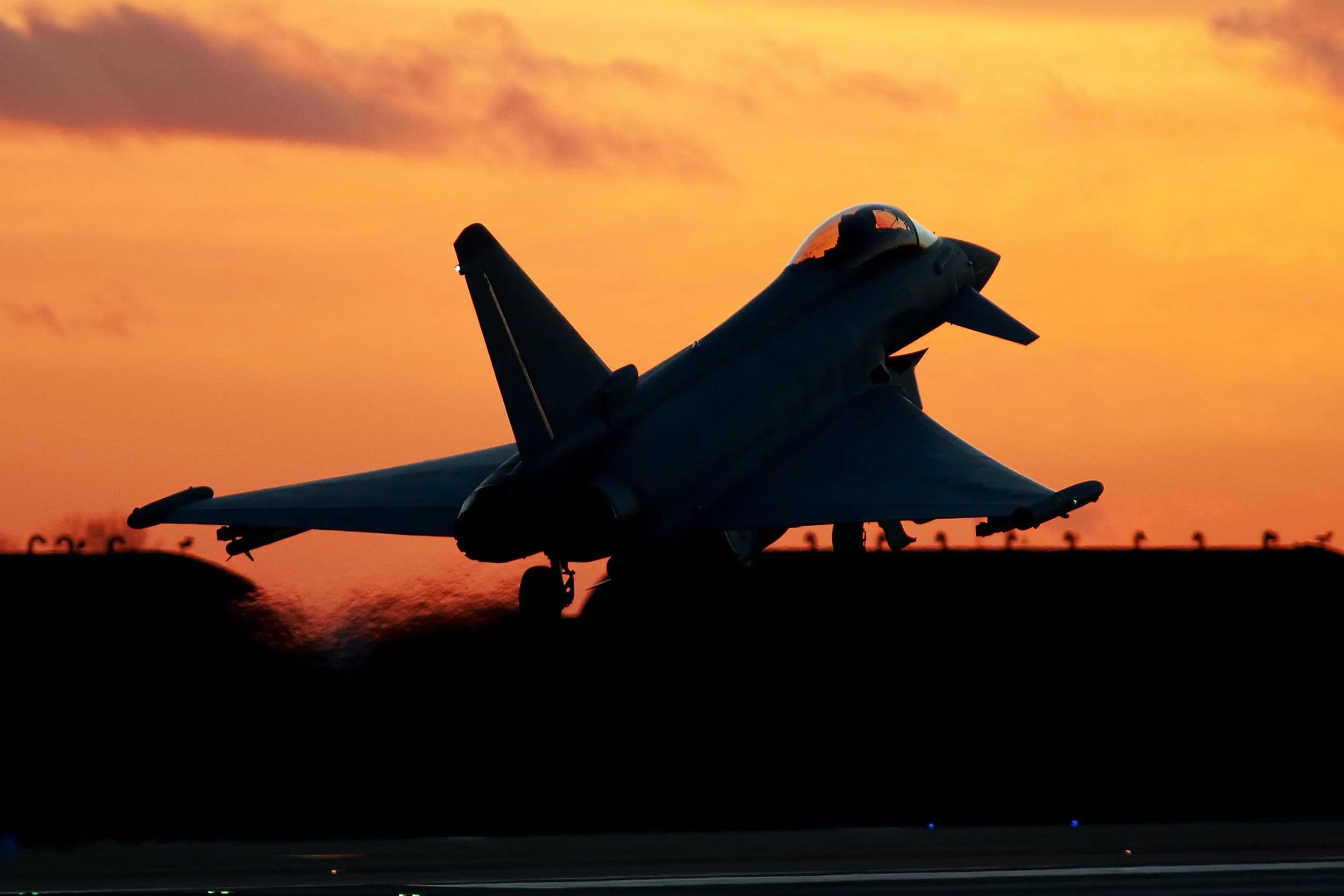 Silhouette of a Eurofighter Typhoon military fighter jet taking off against a dramatic orange sunset.