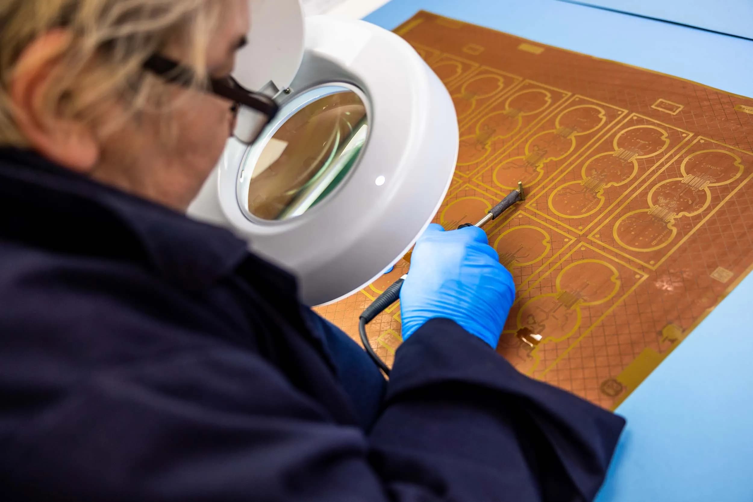 An over-the-shoulder view of an Exception PCB operator looking through a large illuminated magnifier while aligning an orange multi-up flexible circuit panel.