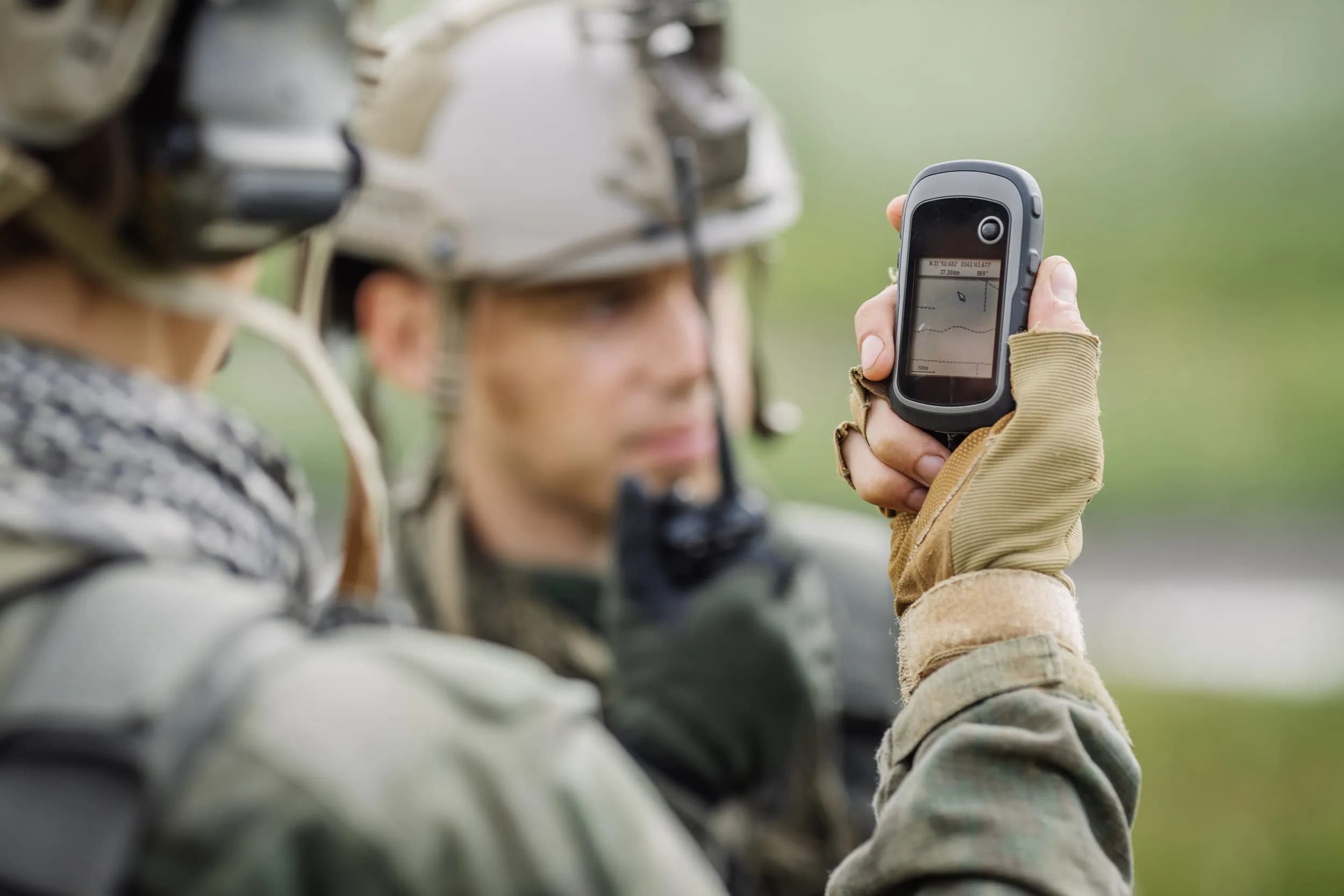 A soldier in tactical gear holding up a ruggedised handheld GPS navigation device with blurred military personnel in the background.