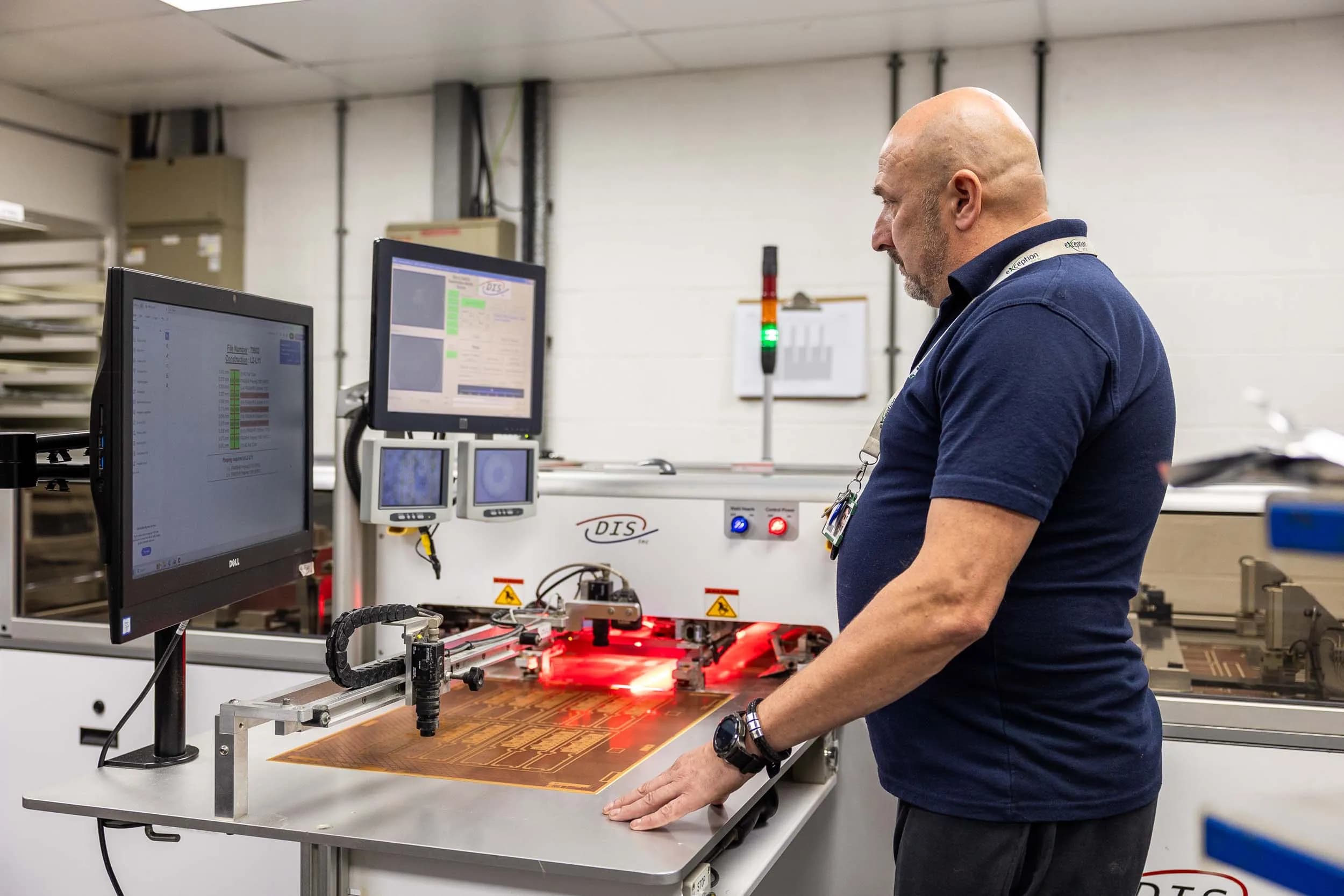 An Exception PCB technician monitoring a multilayer pinless optical alignment workstation under red light.