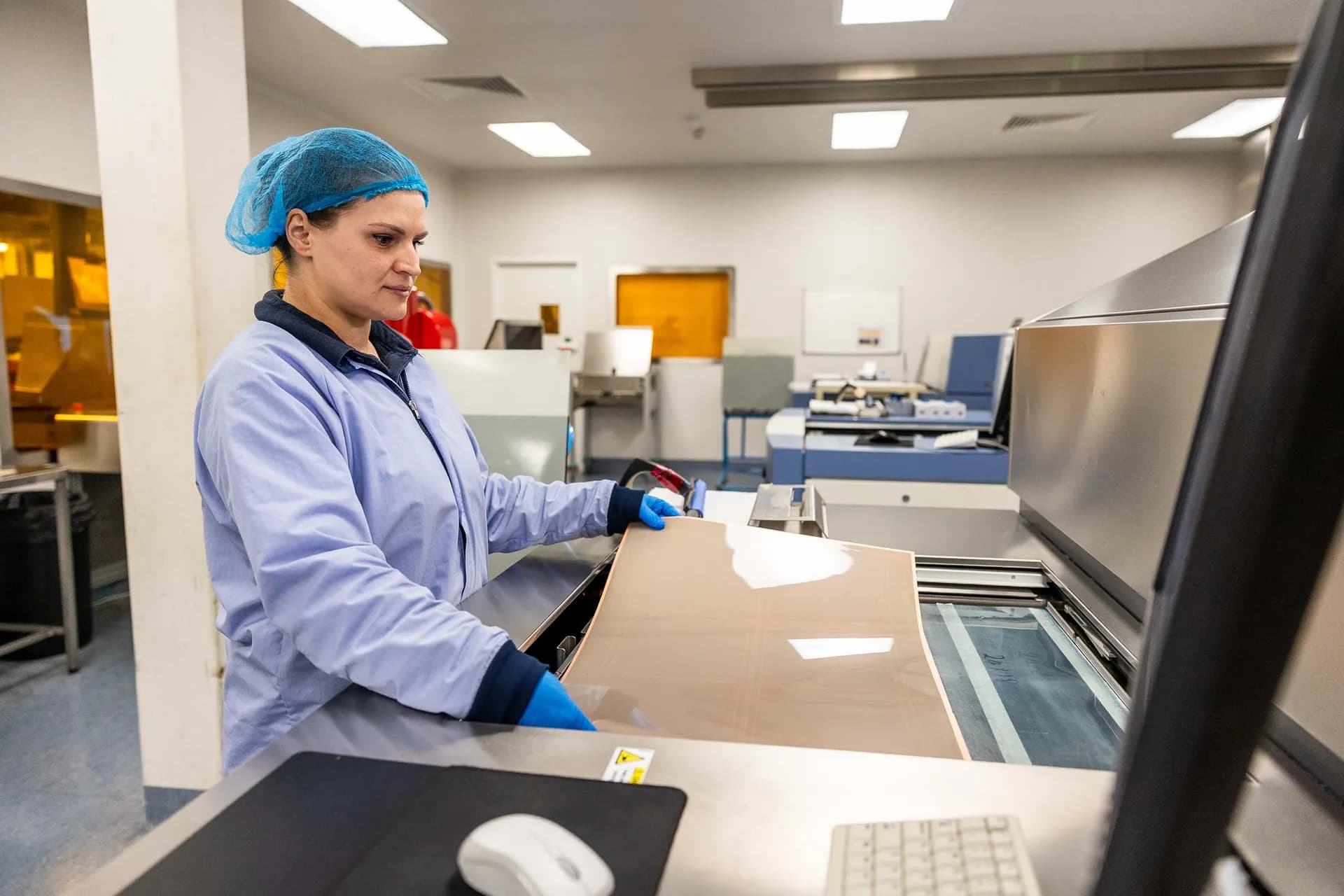 Female Exception PCB technician in blue cleanroom gown and hairnet loading a large bare copper-clad laminate panel into a flatbed direct imaging machine at the Tewkesbury manufacturing facility.