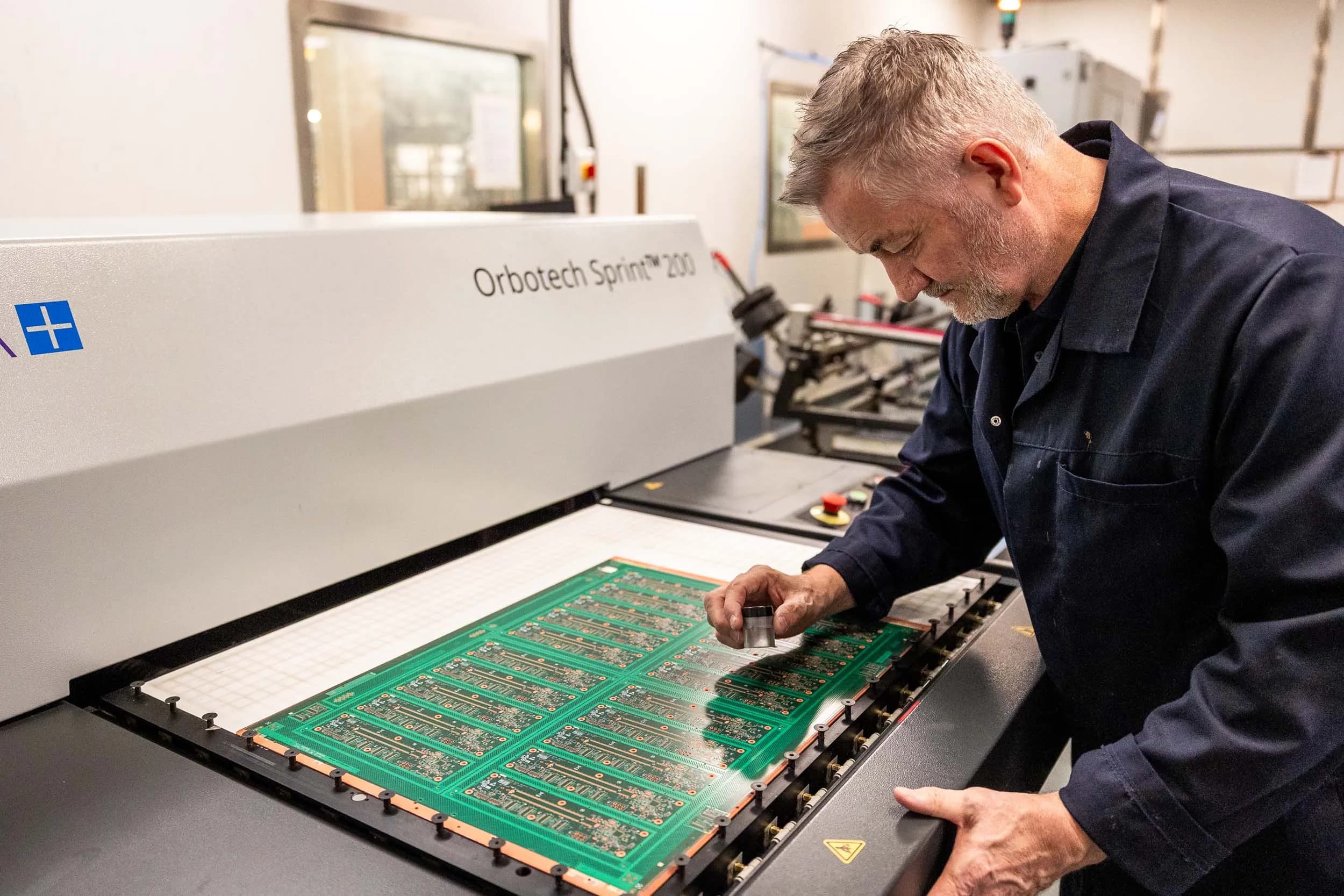 A senior Exception PCB technician in a dark navy lab coat leaning over the scanning bed of an Orbotech Sprint 200 automated optical inspection machine, using a cylindrical loupe to manually inspect a large green multi-up circuit board panel.