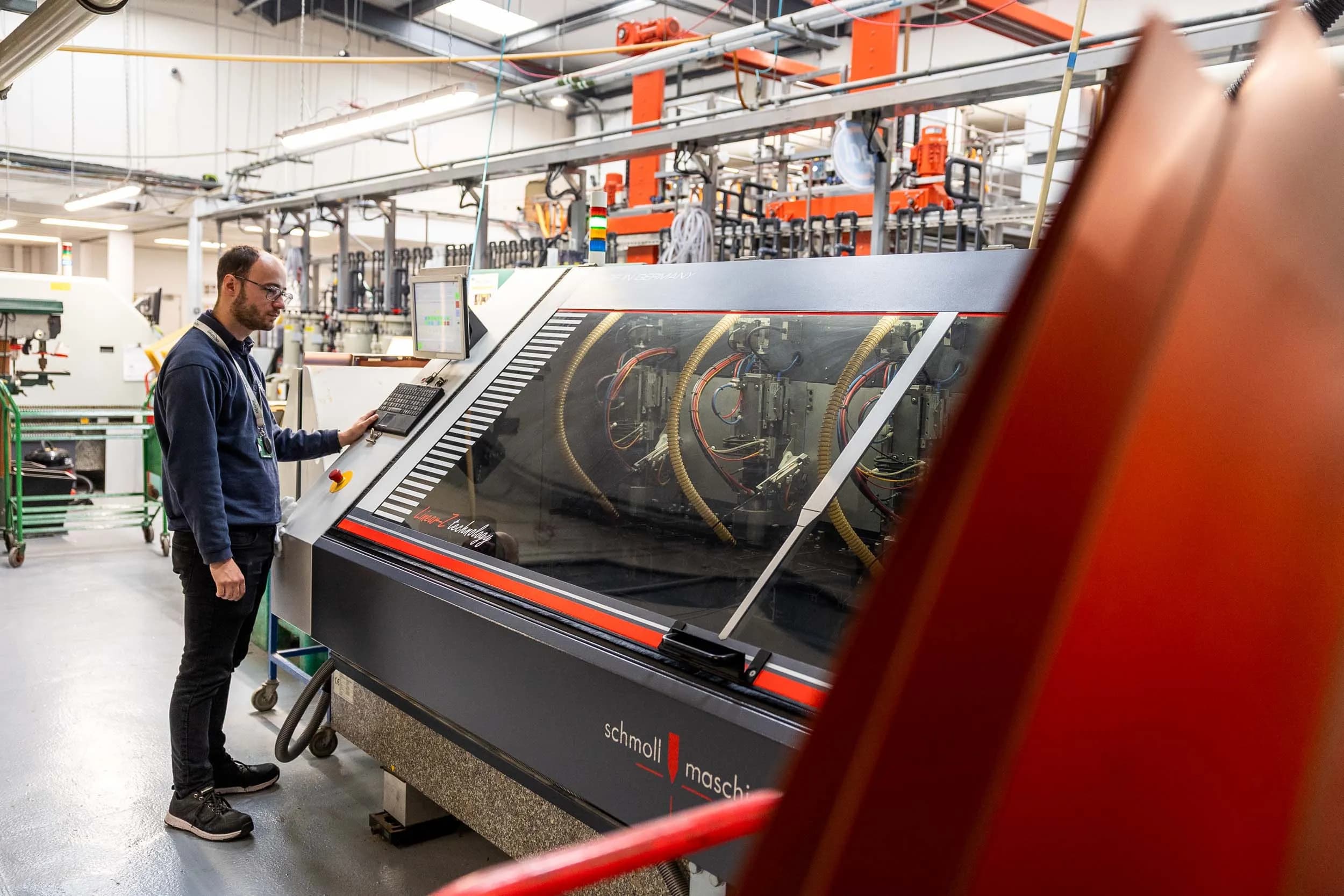 An Exception PCB male engineer in navy fleece and glasses standing at the control panel of a Schmoll Maschinen CNC drilling machine, with multiple drill spindles and flexible extraction hoses visible through the protective glass window.