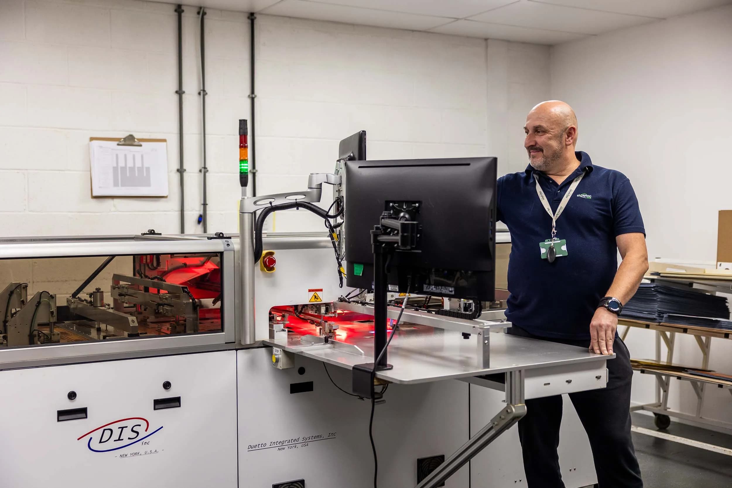 A smiling senior technician standing next to a Duetto Integrated Systems multilayer bonding machine at Exception PCB.