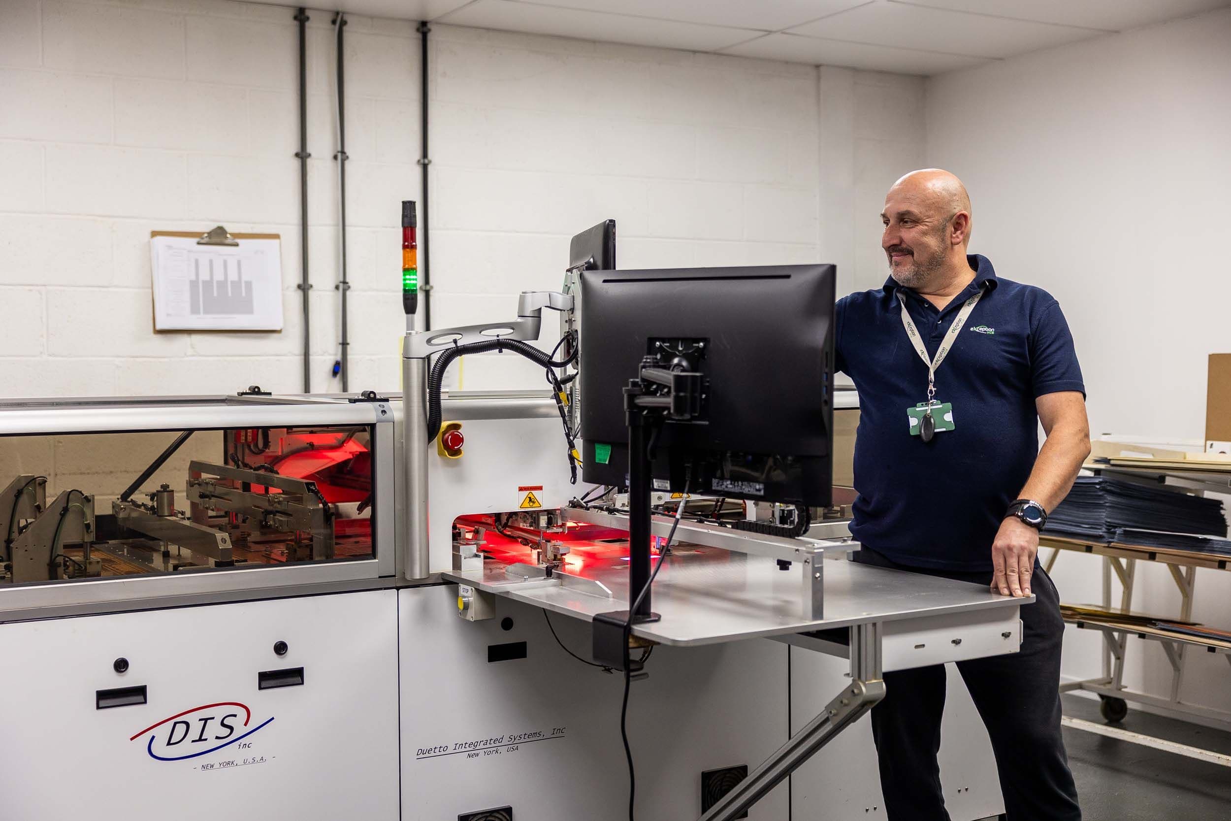 A smiling senior technician standing next to a Duetto Integrated Systems multilayer bonding machine at Exception PCB.