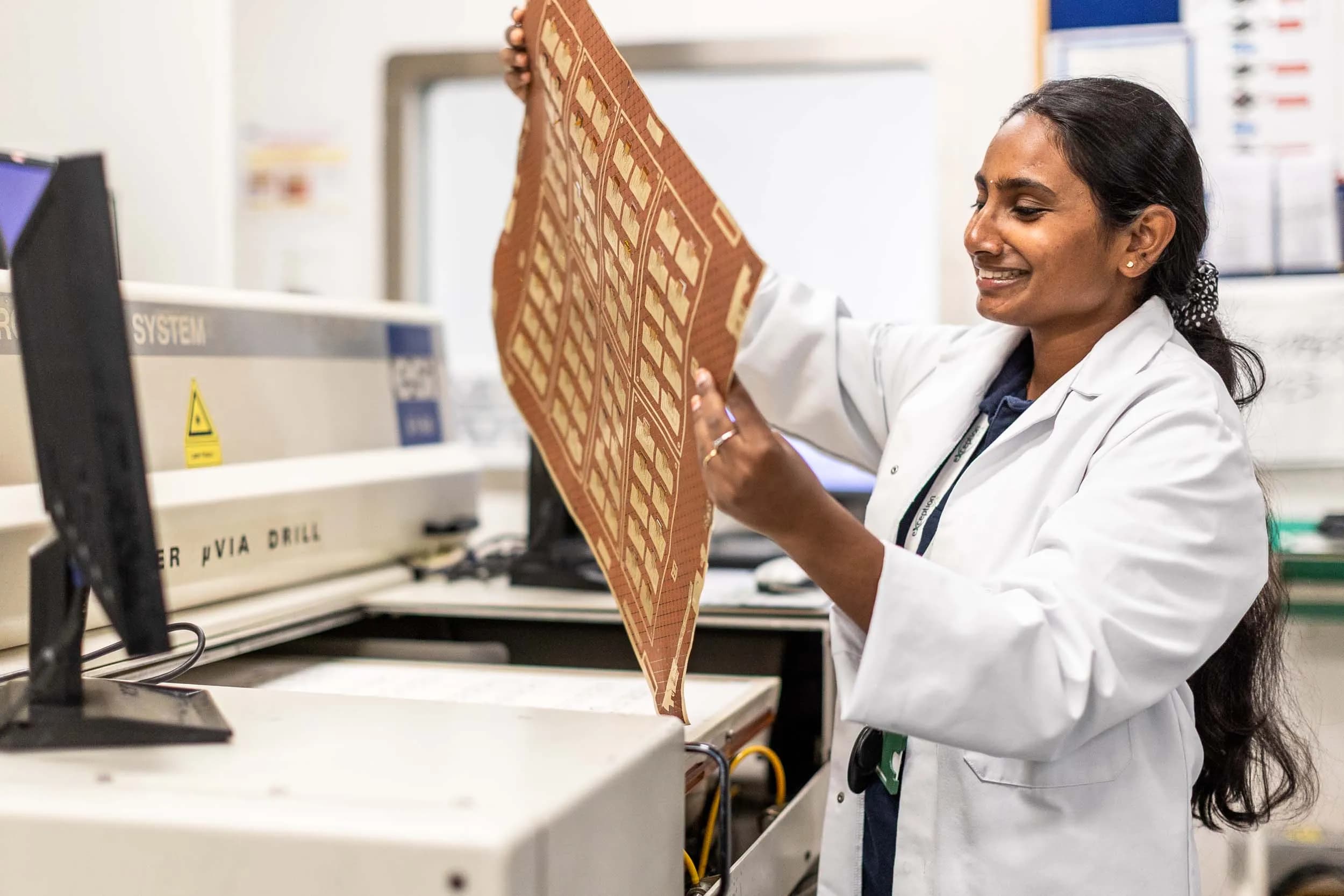Side profile of a smiling Exception PCB engineer holding a flexible circuit sheet next to a micro via drill.