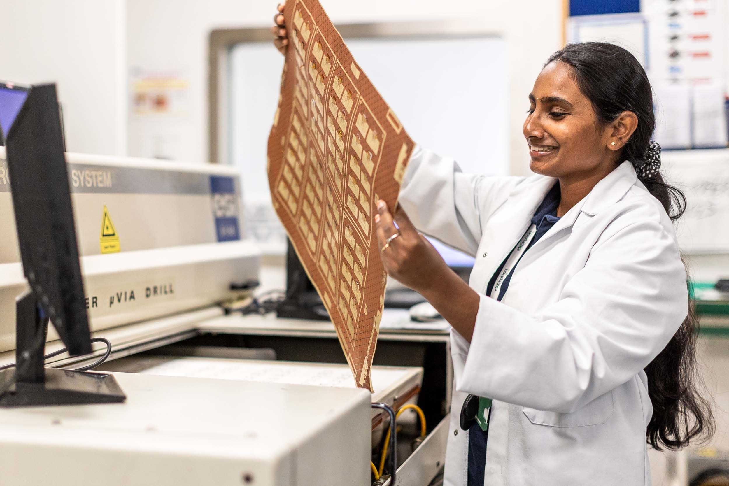 Side profile of a smiling Exception PCB engineer holding a flexible circuit sheet next to a micro via drill.