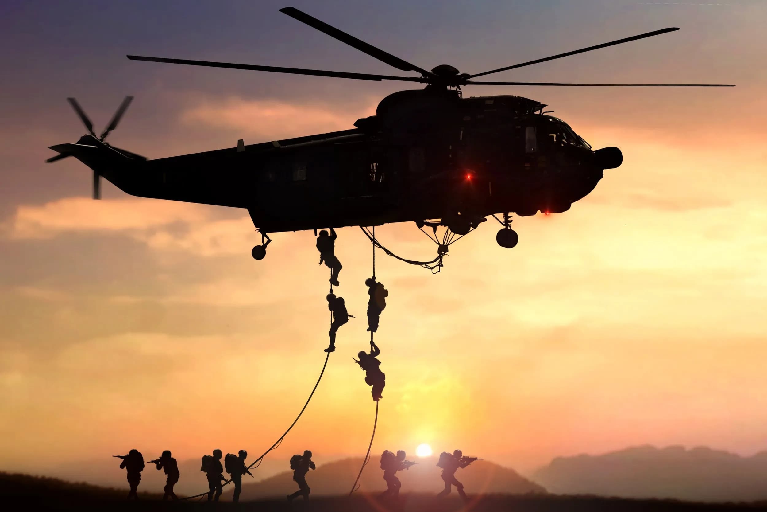 Silhouette of a military helicopter hovering at sunset while tactical soldiers fast-rope down to the ground.