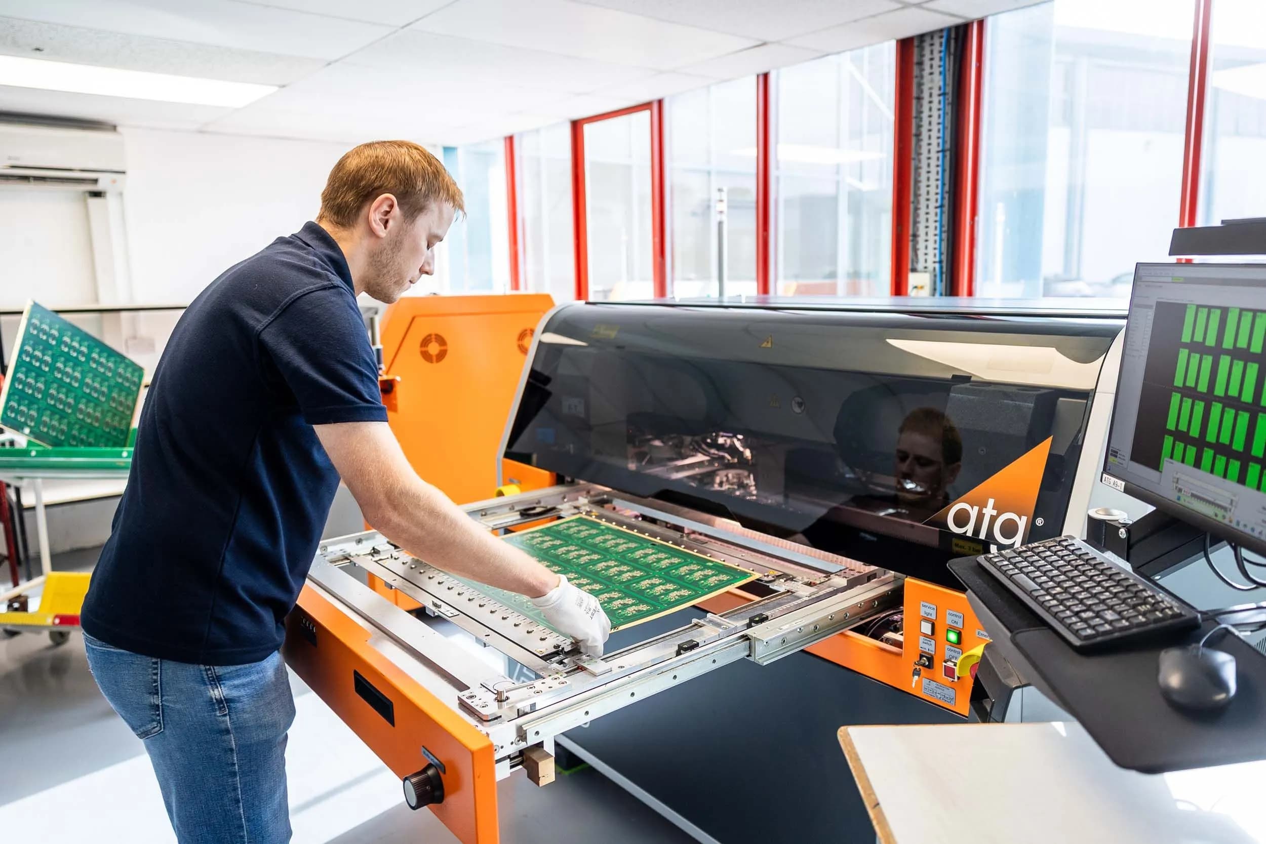 Male PCB technician loading a green solder mask PCB panel into an ATG electrical test machine, portrait orientation showing the full machine height and the bright windowed test room