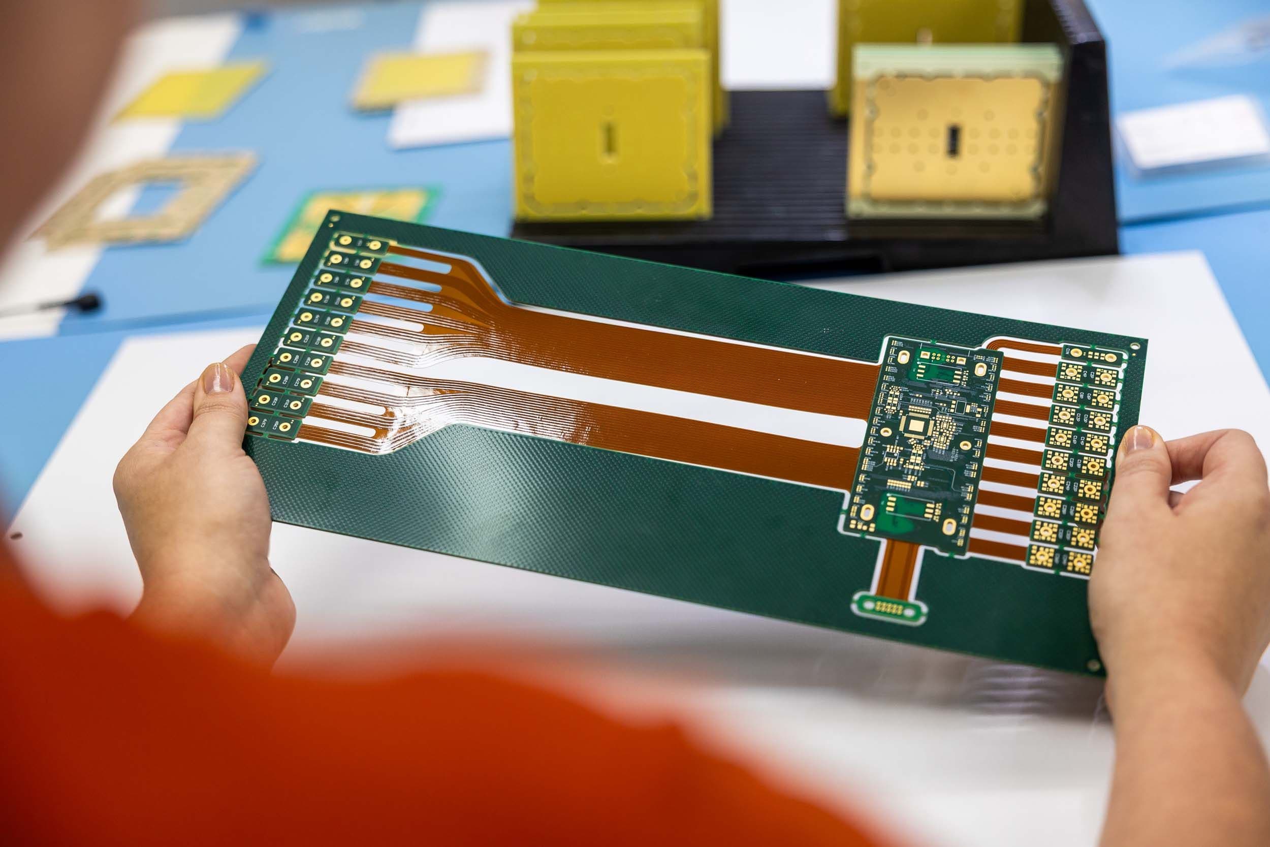 Technician holding a complex green rigid-flex circuit board panel with amber flexible interconnect extensions.