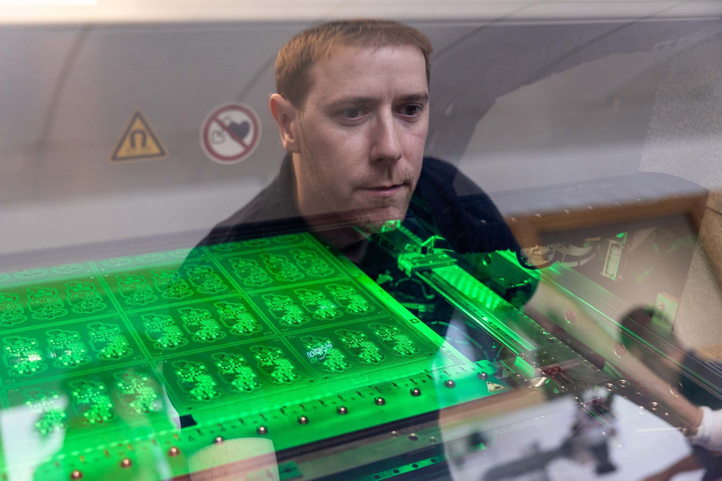 An Exception PCB technician looking through a protective glass window at an illuminated circuit board panel.
