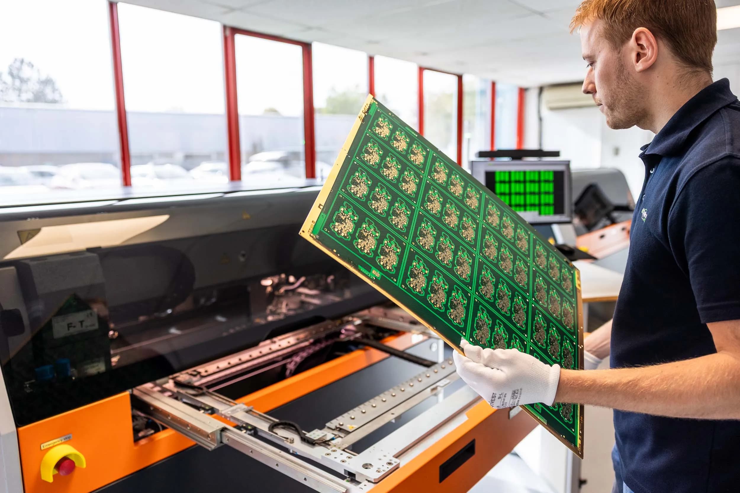 Young male Exception PCB technician in a navy polo shirt and white gloves holding and presenting a large completed green multi-up ENIG printed circuit board panel in a bright production environment.