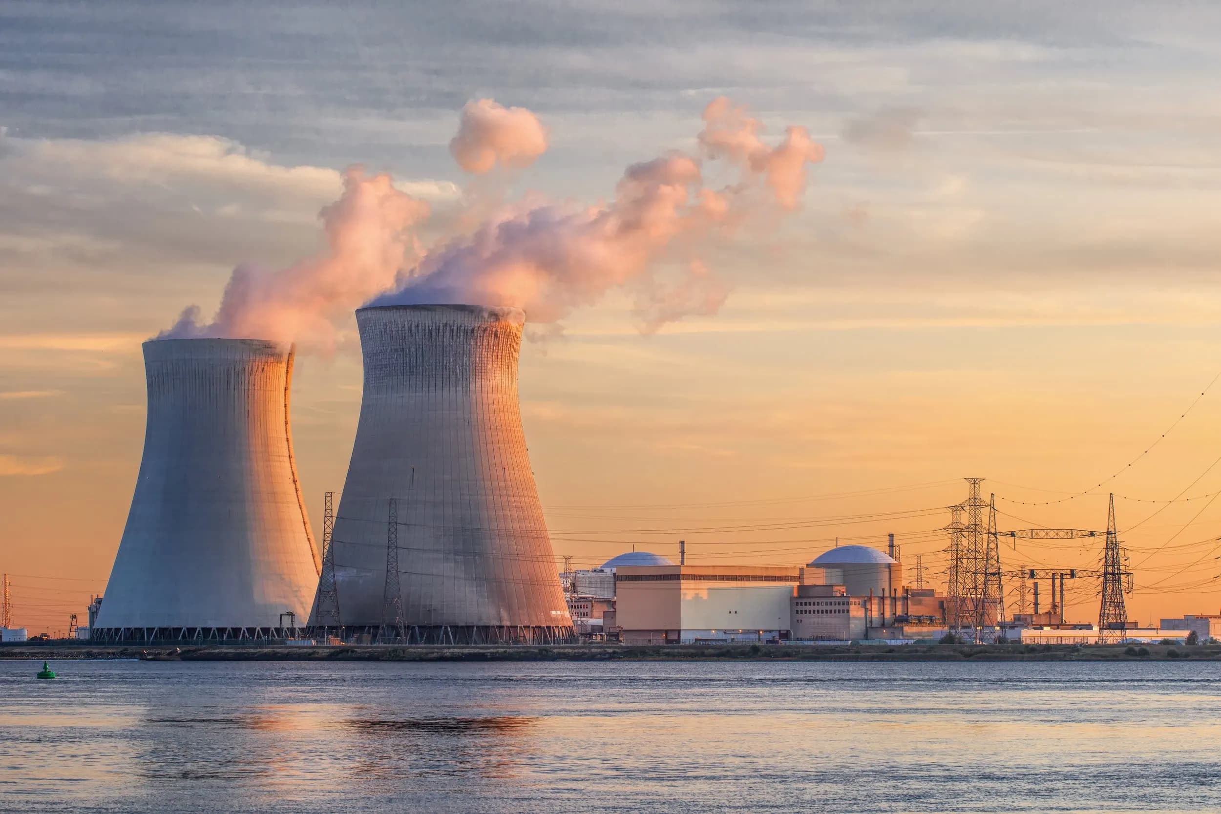 Two large concrete hyperbolic cooling towers of a nuclear power plant emitting steam alongside utility grids and power lines during a warm golden hour sunset.