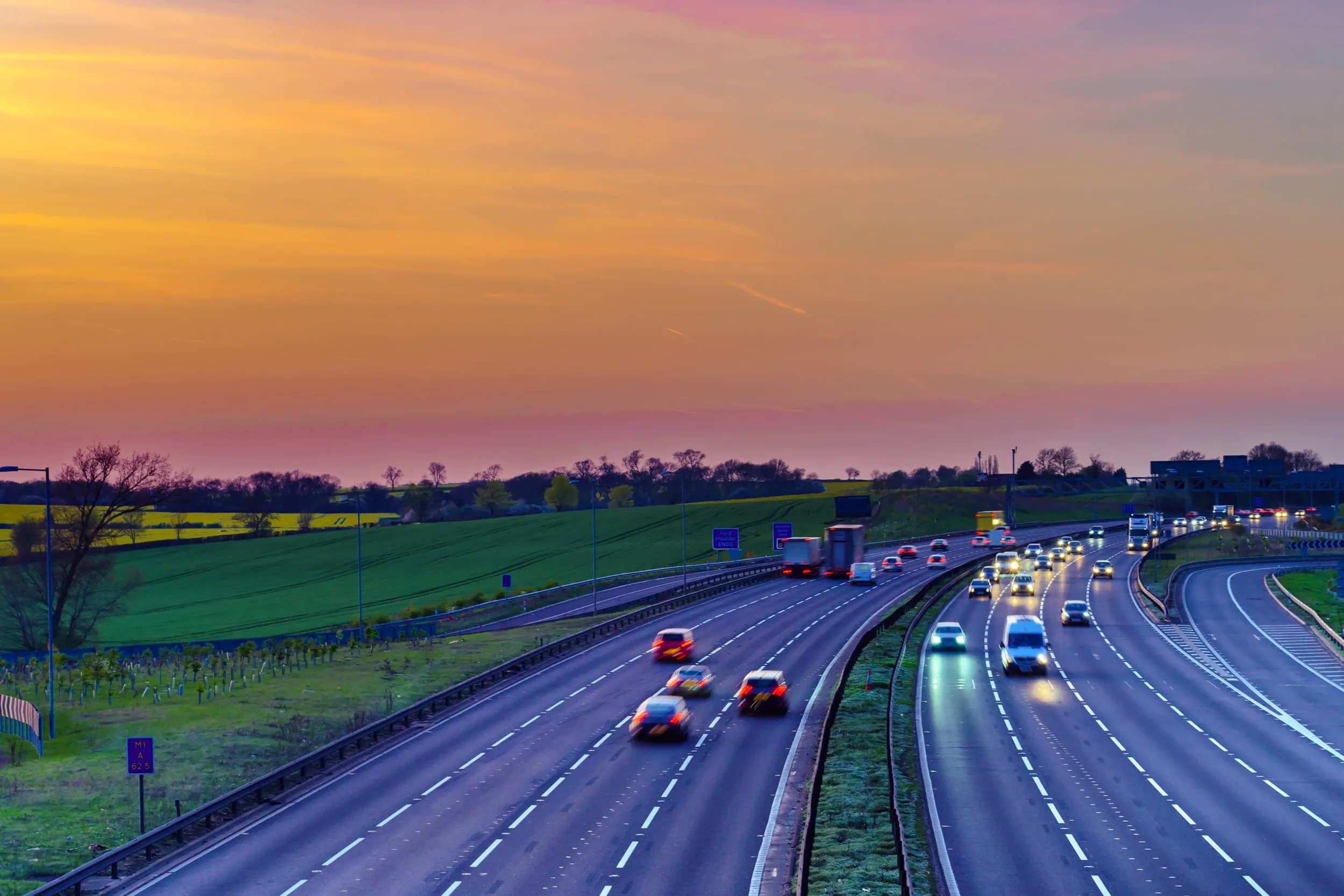 Wide-angle view of a multi-lane UK motorway at sunset with cars and trucks driving under a colourful orange and pink sky next to green fields.